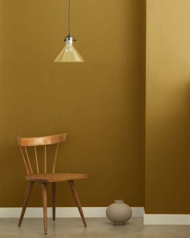 A single-bulb light hangs over a wooden chair and small ceramic pot in front of a wall painted Golden Bark.