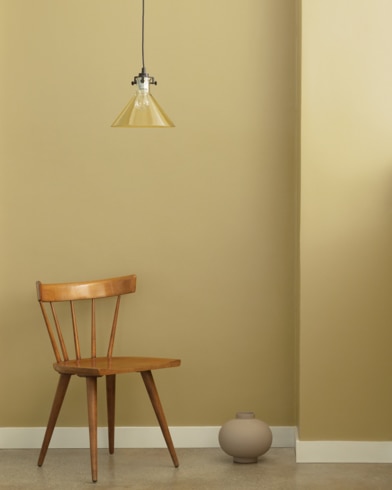 A single-bulb light hangs over a wooden chair and small ceramic pot in front of a wall painted Henderson Buff.