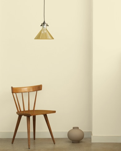 A single-bulb light hangs over a wooden chair and small ceramic pot in front of a wall painted Linen Sand.