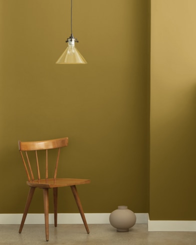 A single-bulb light hangs over a wooden chair and small ceramic pot in front of a wall painted Mustard Olive.