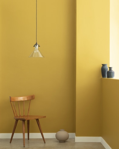 A single-bulb light hangs over a wooden chair and small ceramic pot in front of a wall painted Ochre.