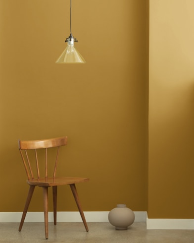 A single-bulb light hangs over a wooden chair and small ceramic pot in front of a wall painted Tapestry Gold.