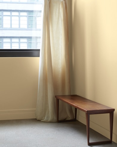 In an Oat Straw-painted room with a wooden bench, a gauzy white curtain hangs next to an open window.
