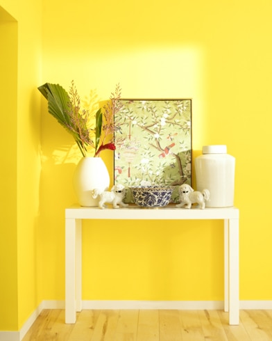 A white table with a collection of white vases, trinkets and abstract art sits against a Bold Yellow-painted wall.