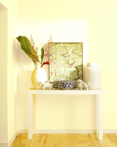 A white table with a collection of white vases, trinkets and abstract art sits against a Light Yellow-painted wall.