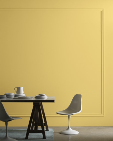 A midcentury modern dining table with two chairs sits in front of a wall painted Goldfield.