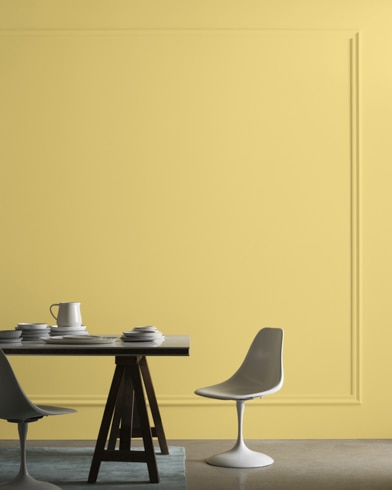 A midcentury modern dining table with two chairs sits in front of a wall painted Laguna Yellow.