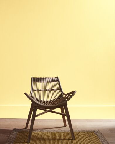 A modern rattan chair with a curved seat sits in front of a Vernazza Yellow-painted wall.