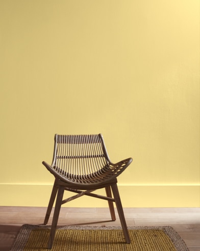 A modern rattan chair with a curved seat sits in front of a Yosemite Yellow-painted wall.