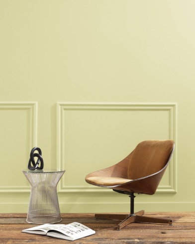 A flat wood riser holds a curved wooden chair, glass vase and open book in front of a paneled wall painted Sesame.