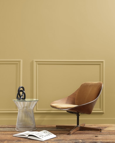 A flat wood riser holds a curved wooden chair, glass vase and open book in front of a paneled wall painted Summerdale Gold.