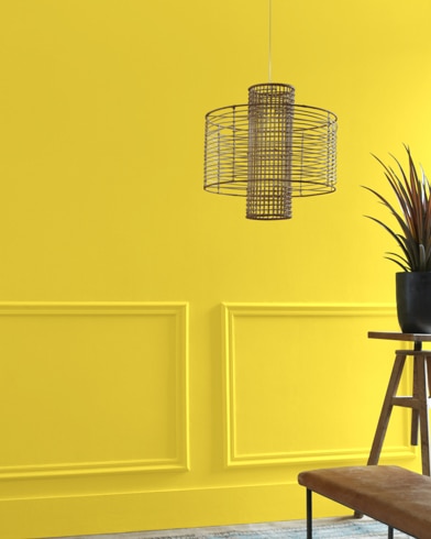 A Yellow Hibiscus-painted living room hosts a wiry light fixture while a wood table and potted plant are just out of focus.