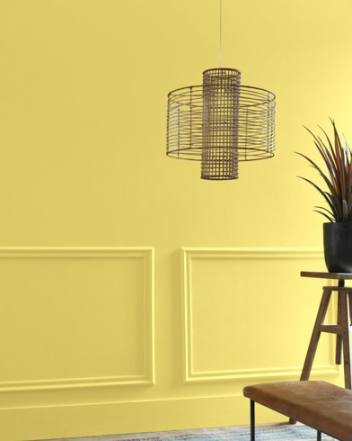 A Yellow Tone-painted living room hosts a wiry light fixture while a wood table and potted plant are just out of focus.