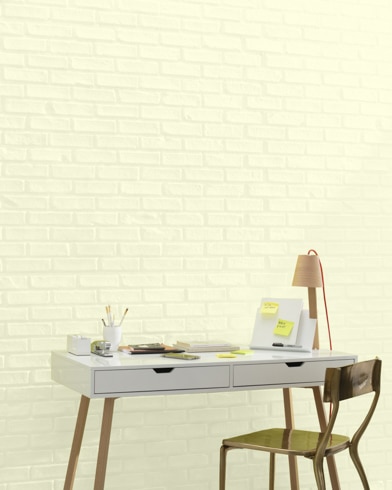 A white and copper writing desk and a bronze metal chair sit in front of a brick wall painted Barely Yellow.