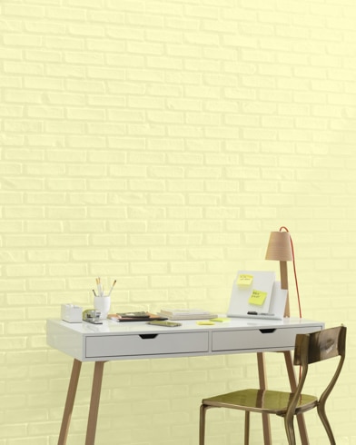 A white and copper writing desk and a bronze metal chair sit in front of a brick wall painted Lemon Glow.