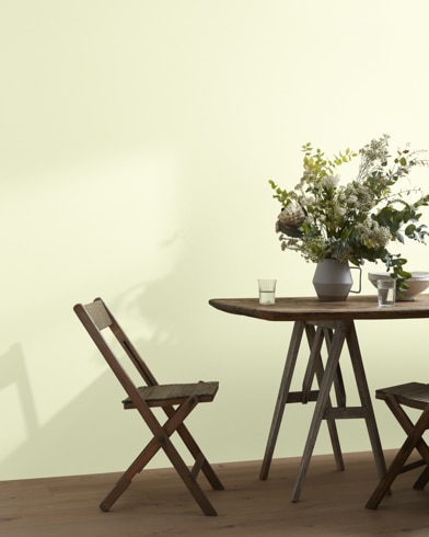 A wood folding chair sits next to a table topped with a greenery-filled vase in front of a Aspen White-painted wall.