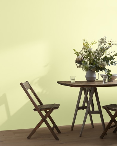 A wood folding chair sits next to a table topped with a greenery-filled vase in front of a Light Daffodil-painted wall.