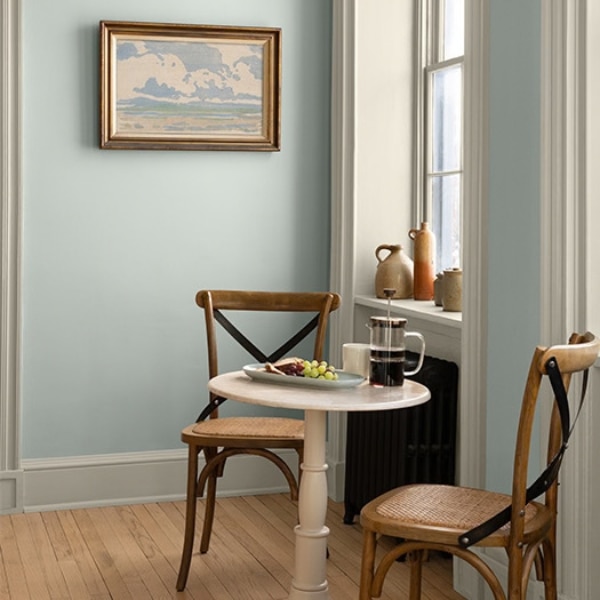 A bistro table sitting in front of a sash window, painted in White Down OC-131 with walls painted in Nelson Blue CW-635, both part of the Colour Trends 2027 collection.